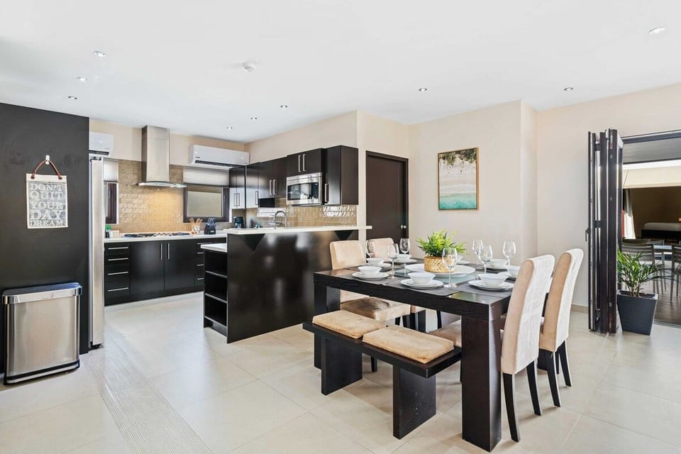 - Dining area with a dark wood table and chairs set for meals, enhancing family and friend gatherings
- Kitchen featuring black cabinets, and a light beige tiled backsplash
- Open layout, connecting the dining area to an outdoor seating space