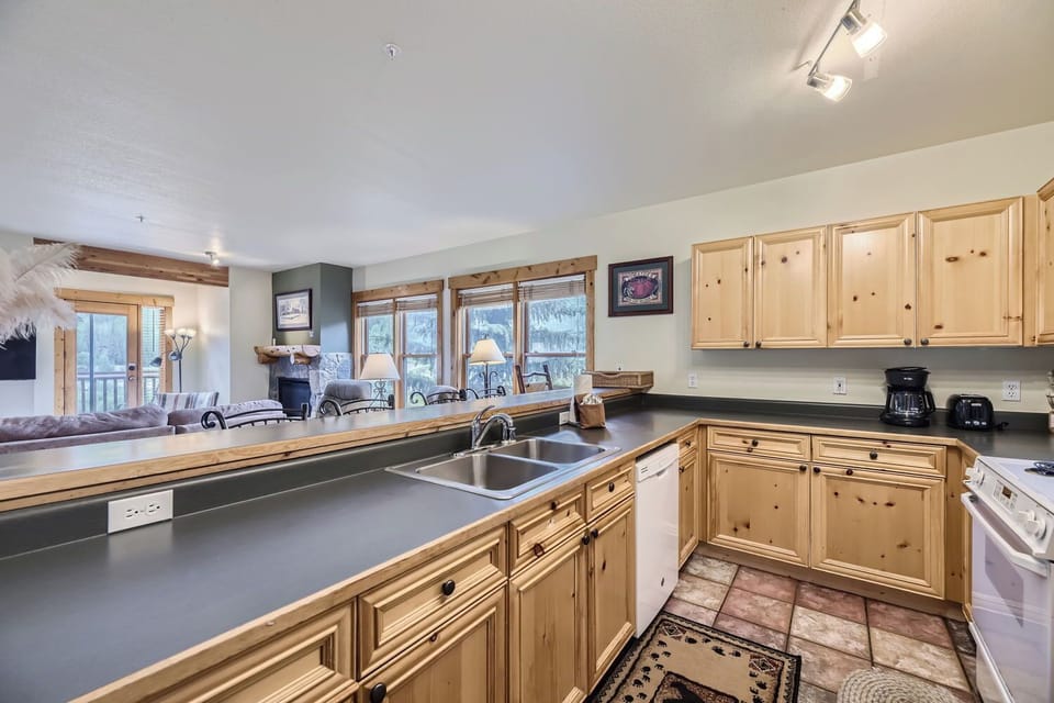 Kitchen with wooden cabinets, a black countertop, and a stainless steel sink. There are appliances like a dishwasher and coffeemaker. The room opens into a living area with large windows.