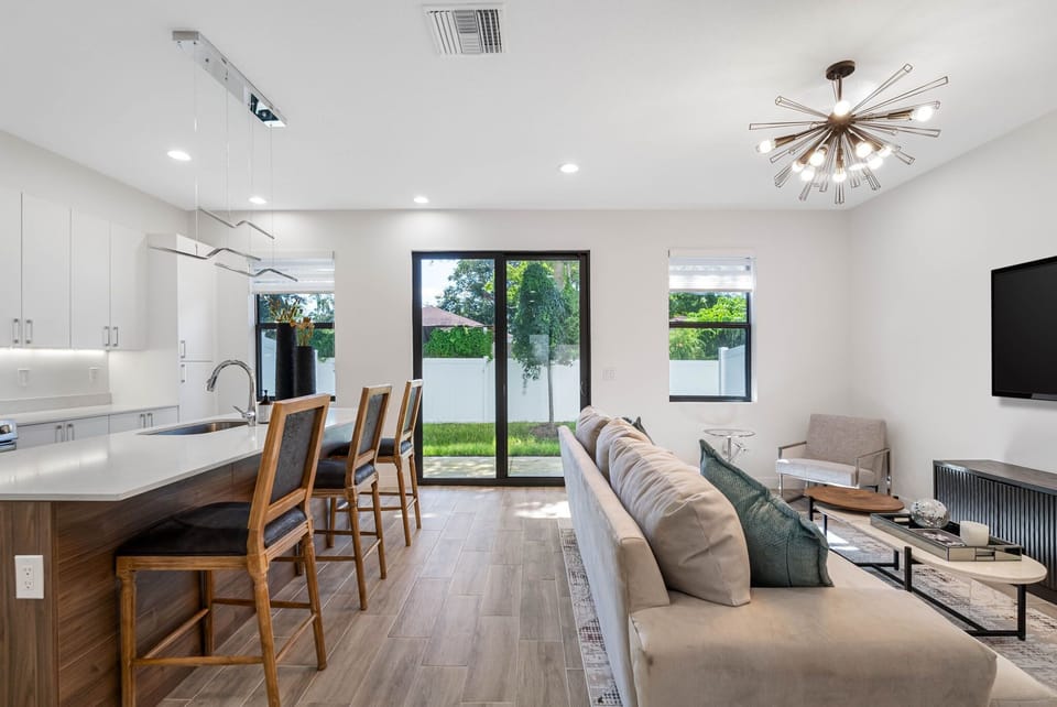 With an open-plan living space, showcasing a sleek kitchen with white cabinetry and enhanced by natural light streaming through expansive windows and glass doors leading to the backyard