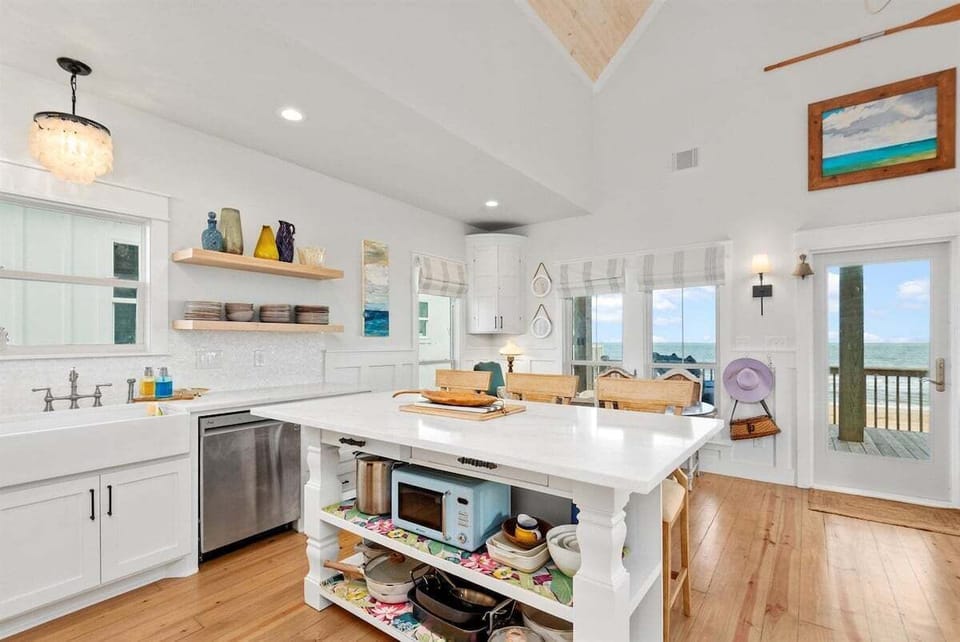 White kitchen with large island, sink, dishwasher, open shelves, wood floors, and ocean-view natural light.