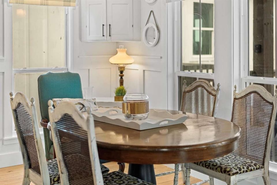 Cozy dining nook with round wood table, upholstered chairs, coastal wall decor, and bright cottage-style windows.