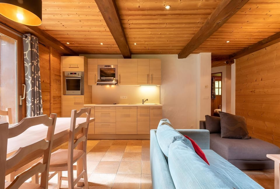 The open-plan living area of the apartment features a comfortable blue sofa with red and brown throw pillows, perfect for relaxing after a day on the slopes. The wooden dining table and chairs provide a warm and inviting space for enjoying meals together,
