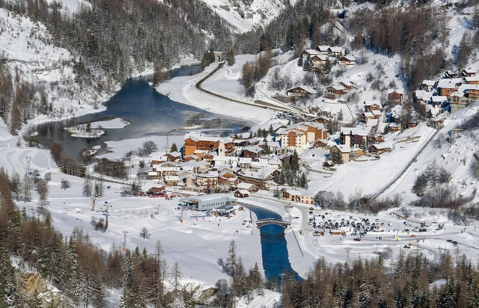 This aerial view showcases the picturesque setting of Tignes-les-Brevières, with the chalet nestled amidst a charming alpine village. The snow-covered landscape, a frozen lake, and a winding river create a breathtaking winter wonderland. The proximity to 