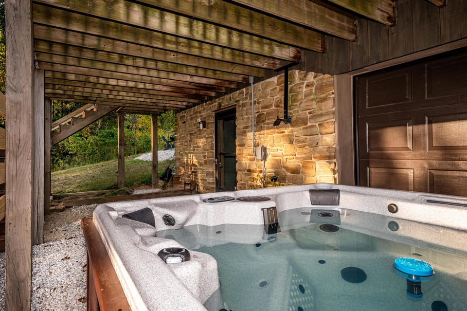 Under the wooden deck, a filled hot tub rests beside a stone wall and brown garage door.