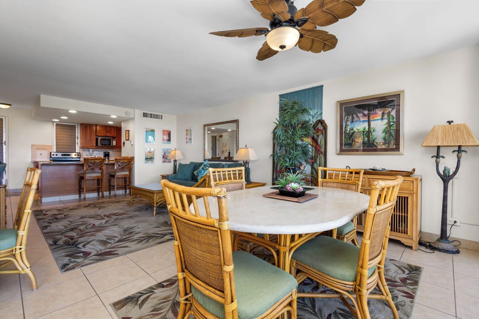 An airy, tropical-themed retreat with island-inspired furniture flows from your dining area to the open kitchen, all cooled by a charming leaf-blade ceiling fan overhead.