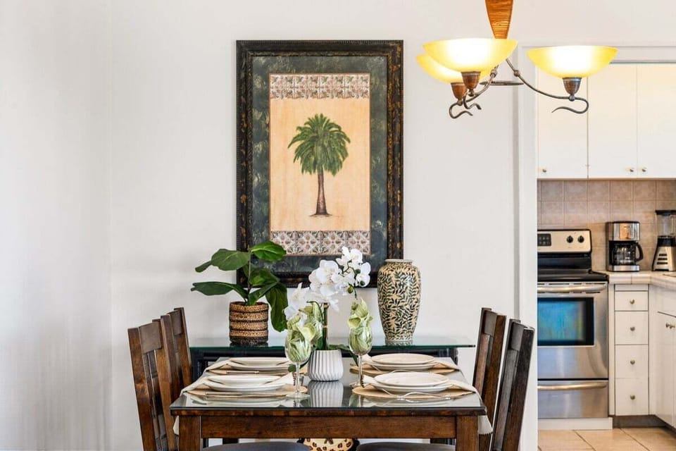 Stylish dining nook featuring a glass‑top table, tropical wall art, warm wood flooring, and soft lighting for relaxed indoor meals.