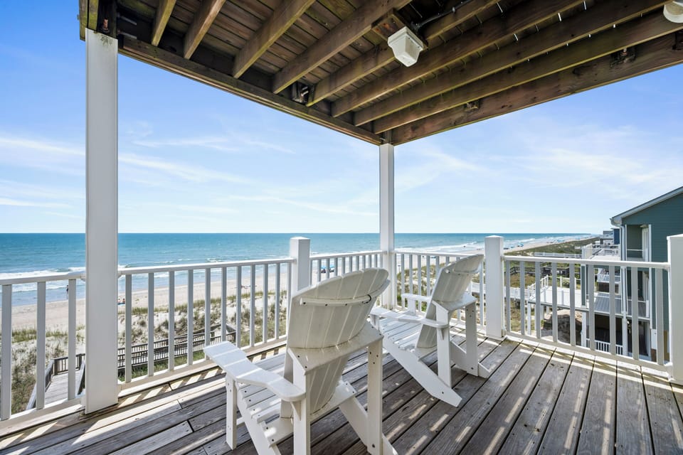 A covered oceanfront deck with white Adirondack chairs showcasing direct beach access and panoramic ocean views, framed by dunes and coastal vegetation.