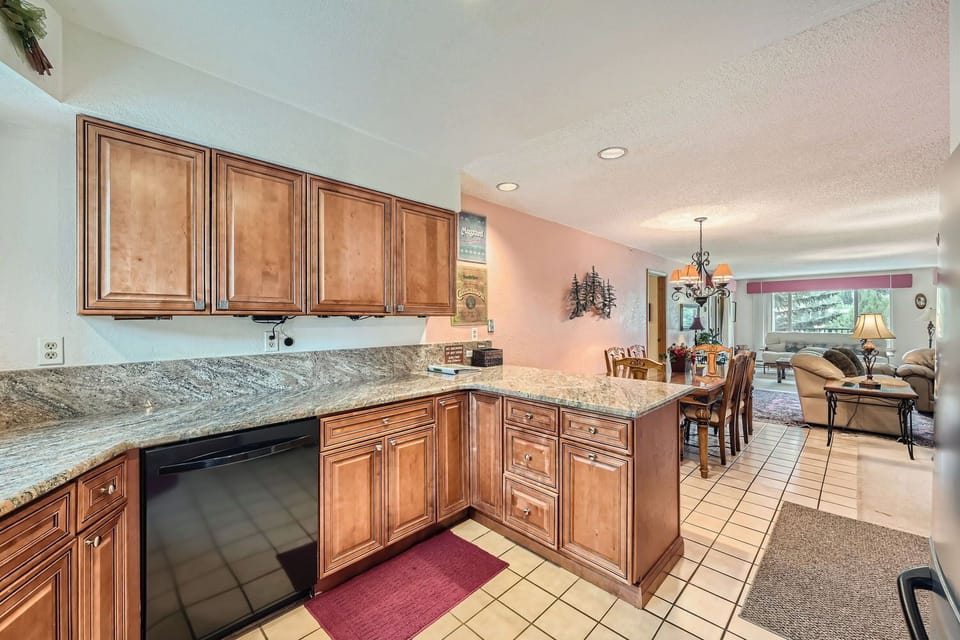 A kitchen with wooden cabinets, granite countertops, and a black dishwasher. Adjacent to the kitchen is an open dining area with a long table and a living room with seating and large windows.
