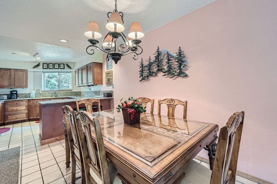 A dining room with a wooden table and six chairs. A chandelier hangs above, and a decorative metal tree art piece adorns the wall. The kitchen is visible in the background.