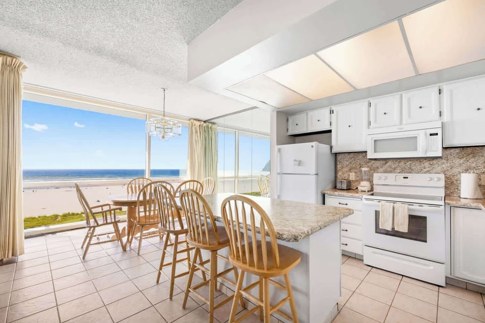 Oceanfront cooking and dining with a view! This fully equipped kitchen features an island with bar stools, a dining table, and expansive windows showcasing stunning beach scenery
