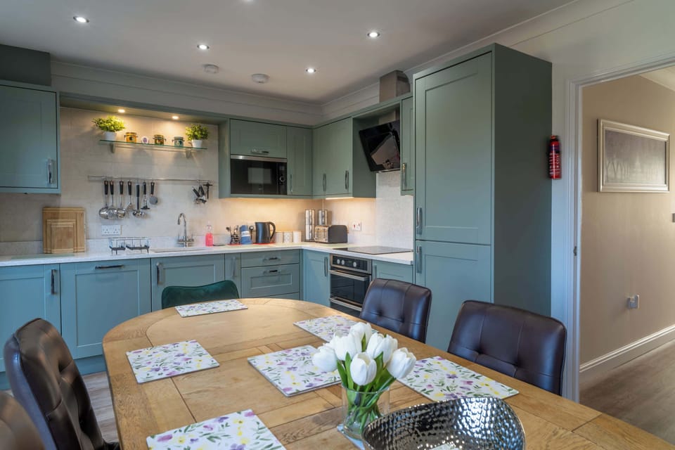 An open-plan kitchen and dining area with sage green cabinetry, integrated appliances, and clever shelving. The rustic wooden dining table complements the modern design, set with floral placemats and tulips in a glass vase.