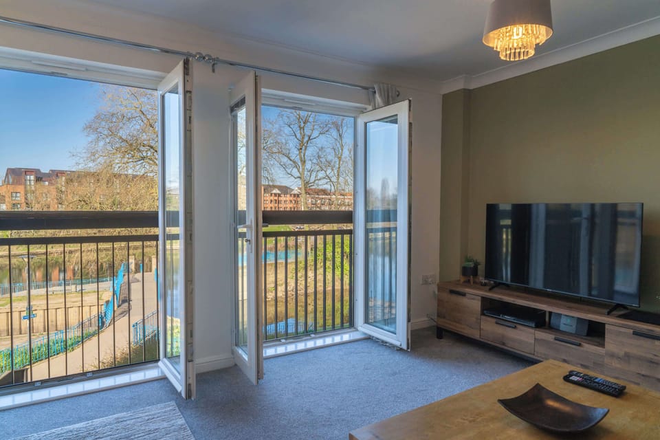 A bright and airy living area with large French doors that open onto a scenic riverside view. Natural light floods the space, highlighting the modern media console and smart layout that perfectly blends comfort and functionality.