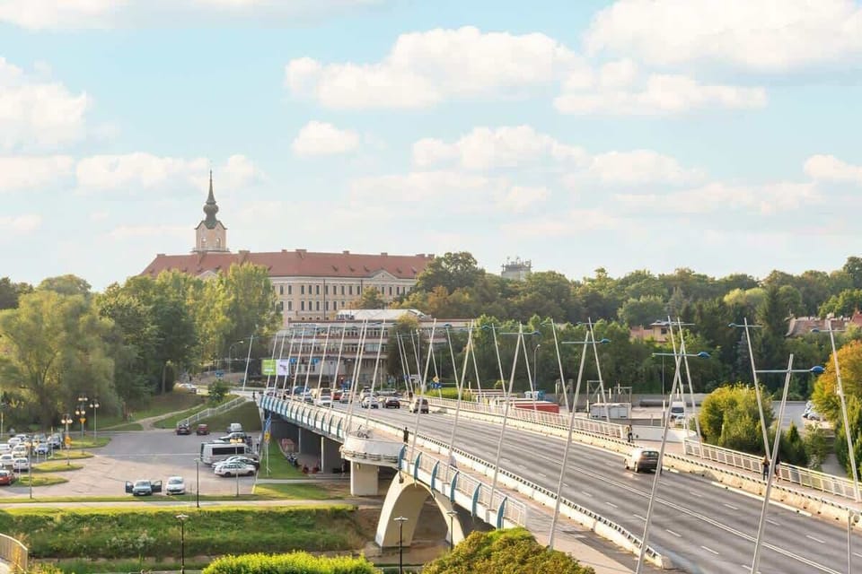 A scenic cityscape with a bridge over a river and historic architecture in the background. Greenery surrounds the area.