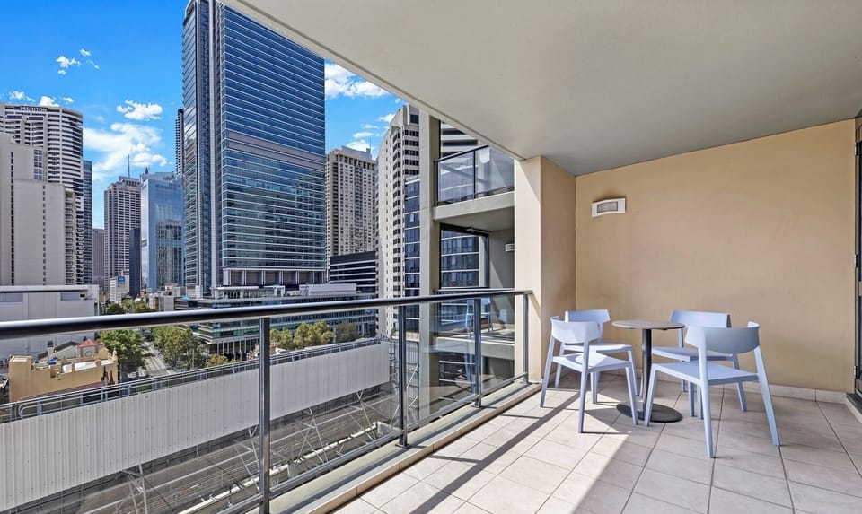 An outdoor balcony area with glass railings, furnished with a patio table and chairs, overlooking modern city high-rises.