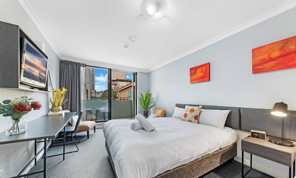 A bright bedroom showcasing a neatly made double bed with white linens, decorative orange pillows, bedside tables with lamps, and a wall-mounted TV.