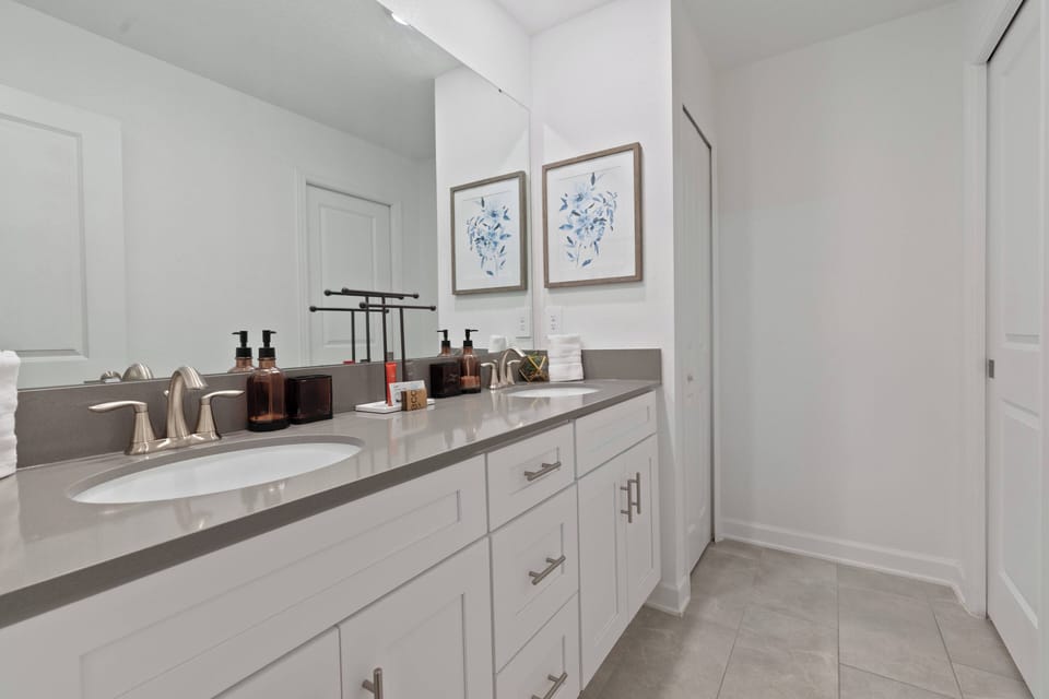 Experience a bright and airy bathroom with stylish white cabinetry and a spacious double vanity topped with polished stone. Delicate artwork adds charm, and neutral tile flooring enhances the serene ambiance.