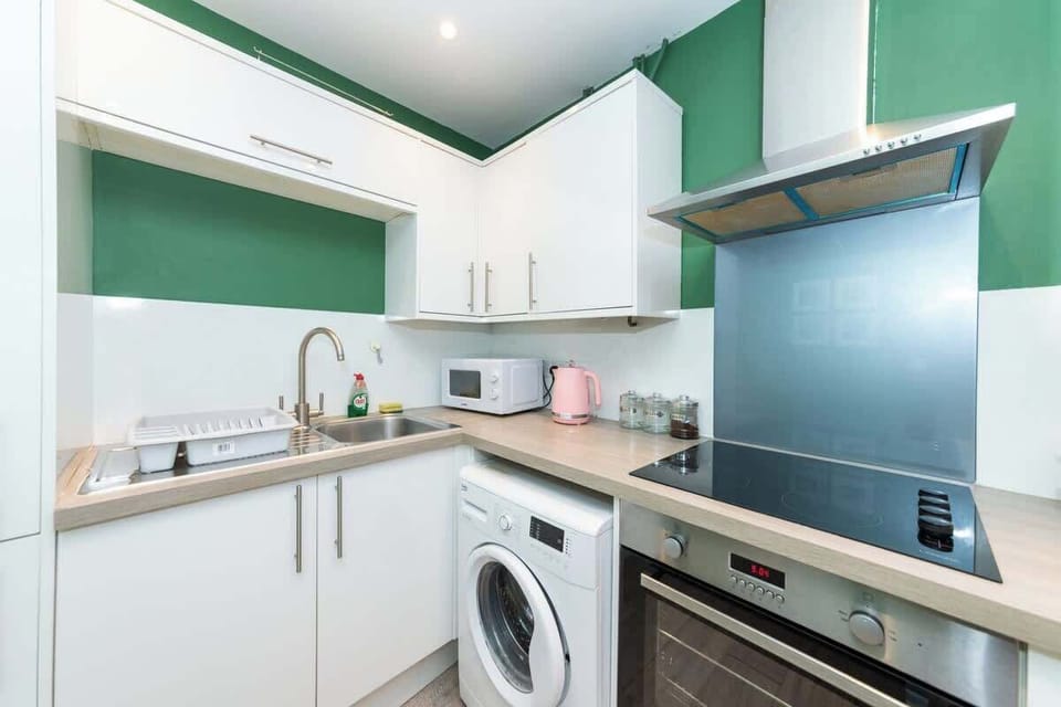 Modern kitchen with green walls, white cabinets, a washing machine, electric stove with oven, stainless steel hood, microwave, and pink appliances on a light wood floor.