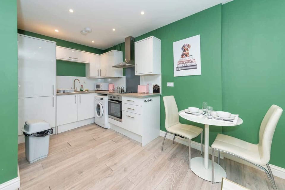 Modern kitchen with white cabinets and appliances, green walls, and light wood flooring. A small round dining table with two cream chairs is set for two. A dog-themed artwork hangs on the wall above the table.