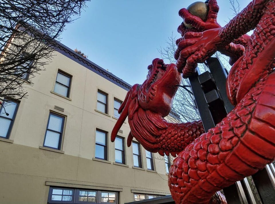 Glossy red dragon sculpture with detailed scales and flowing mane framed by bare tree branches against a pale building facade.