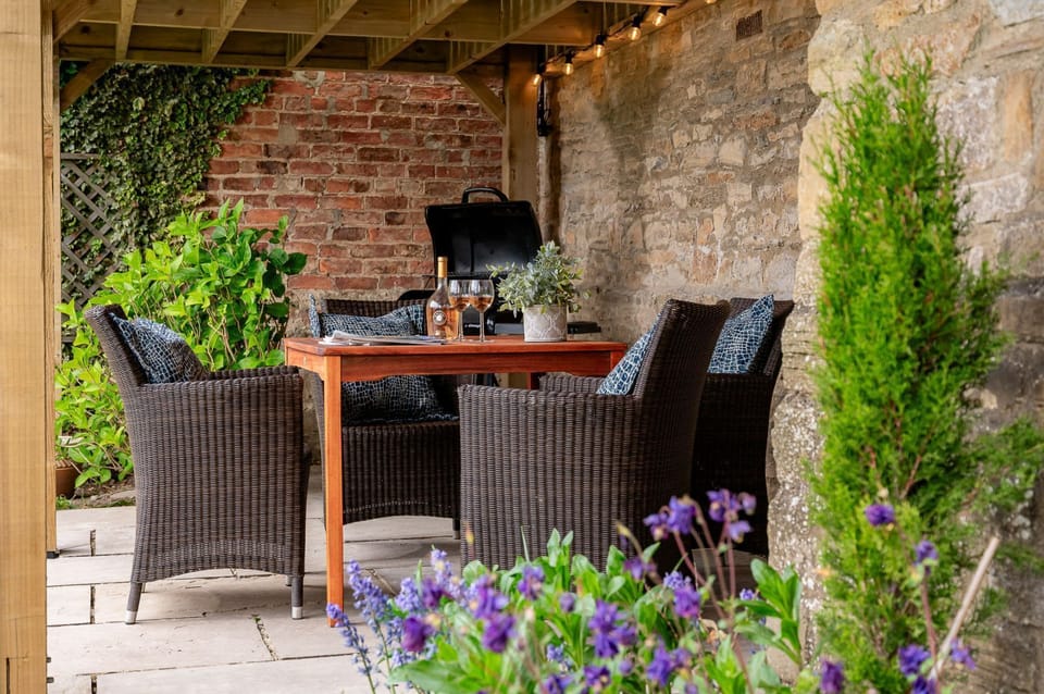 Maison Cottage covered patio dining area with wicker seating, BBQ and rustic stone wall backdrop