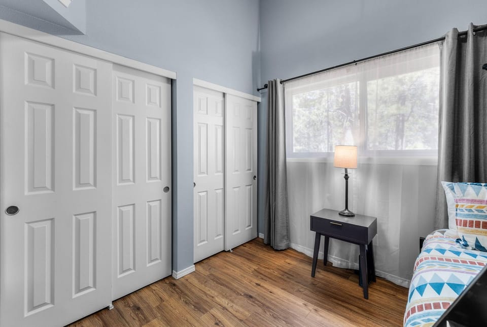 Soft blue walls and warm wood floors create a restful retreat with natural light filtering through sheer curtains, highlighting the clean lines and generous closet space.