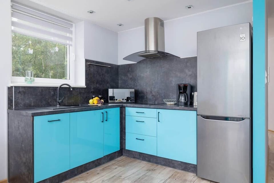 A bright and practical kitchen features turquoise cabinetry, built-in appliances, and a wide window overlooking greenery.