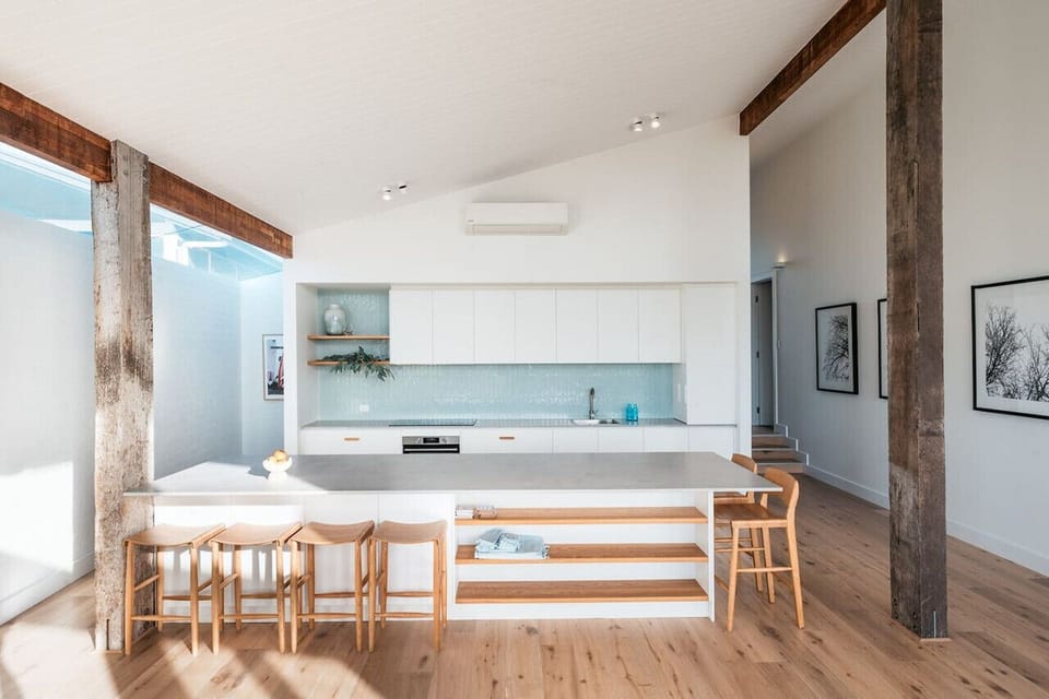 A modern kitchen with a sleek design, featuring white cabinetry, a light-colored countertop, and wooden stools. The space has high ceilings with exposed wooden beams and columns, adding a rustic touch. The kitchen is part of an open-plan area with wooden 