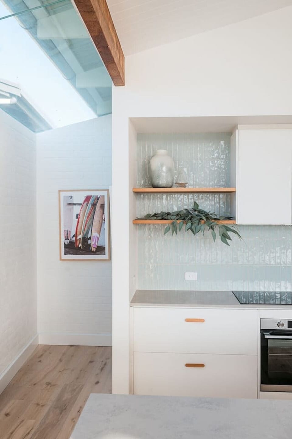 The backsplash is adorned with light blue tiles, and there are wooden shelves holding decorative items like a vase and some eucalyptus leaves. A framed picture of surfboards hangs on the wall, adding a touch of character. The kitchen has a sleek design wi