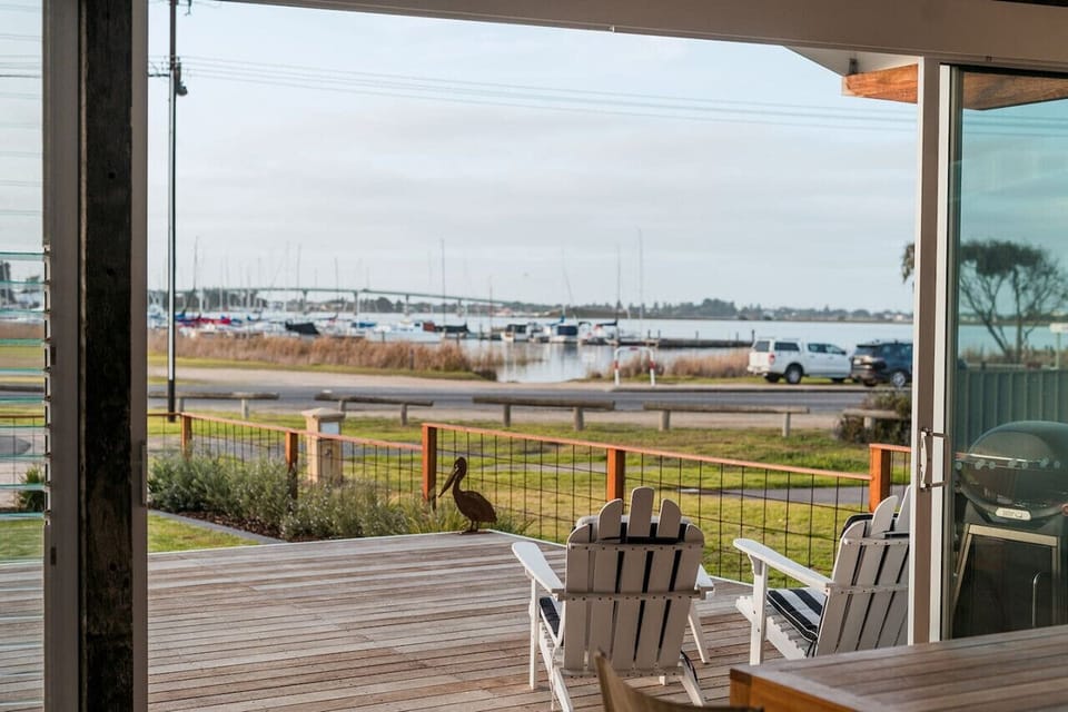 A wooden deck with two white Adirondack chairs offers a view of a harbor with boats and a bridge in the background, and a pelican is standing on the deck.