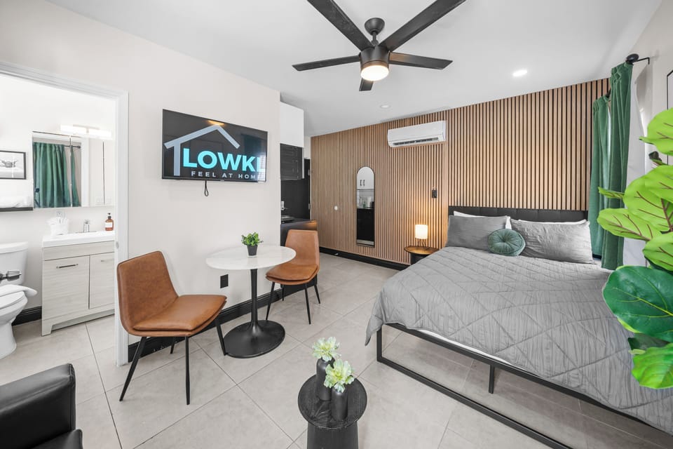 This inviting studio features a cozy queen-size bed, a comfortable leather sofa bed, and stylish decor with vibrant wall art and greenery. Natural light from large windows and a wood-accented wall create a warm, relaxing atmosphere.
