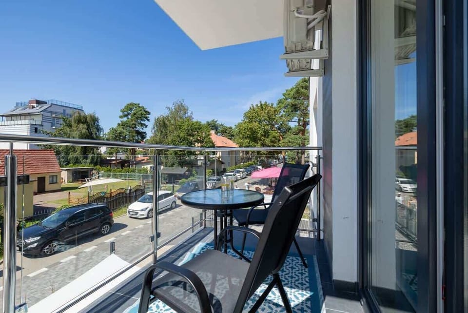 A sunny balcony with a small table and chairs, overlooking a quiet neighborhood with green trees and neatly maintained streets.