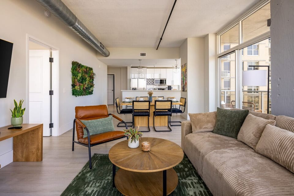 Open-concept living and dining area with a beige sofa, leather chair, 65` TV and wooden coffee table. The dining space features a six-seater table with a view of the kitchen. Bright and airy, with floor-to-ceiling windows and vibrant wall decor.