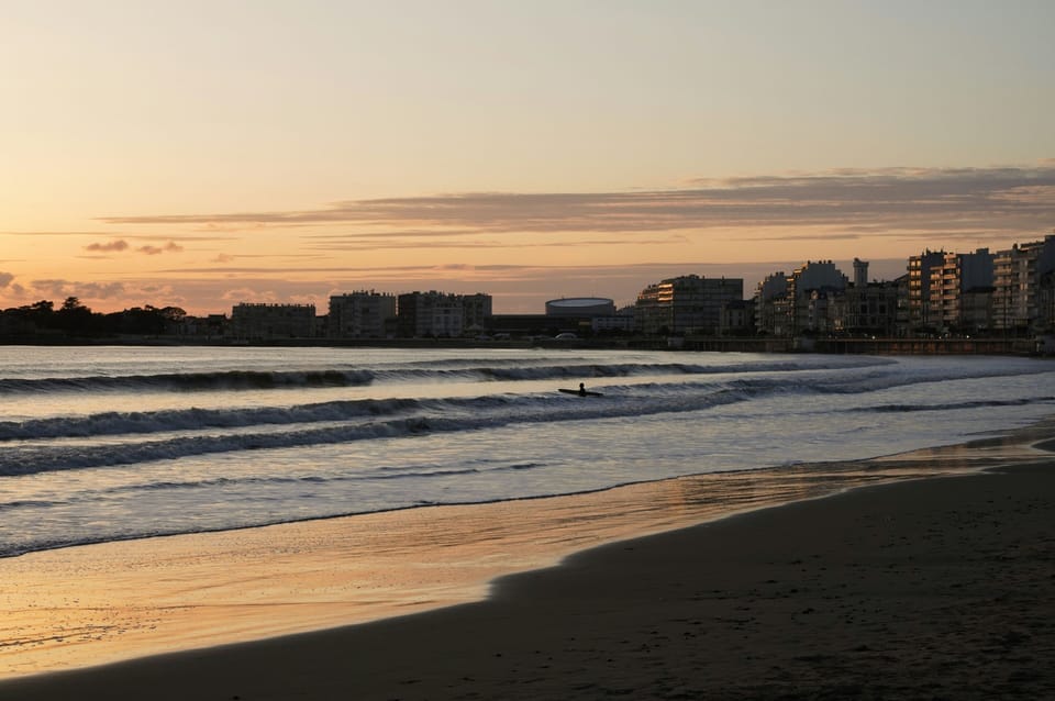 Wide sandy beach at sunset with gentle surf and town buildings silhouetted along the shoreline