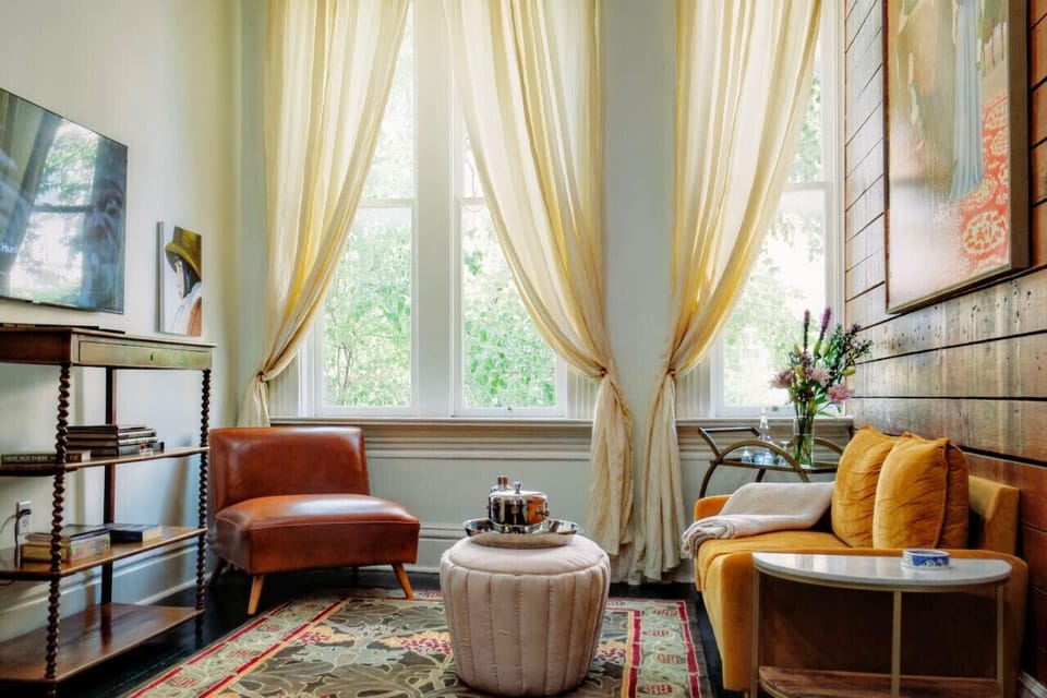 Light filled living room overlooking Main Street with original artwork, original shiplap, flat screen streaming tv, books, silk curtains.