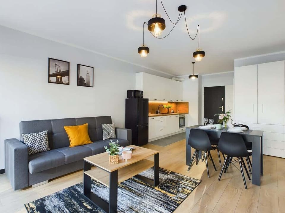 A wide living room view with a dining table, black sofa, and windows bringing in natural light.