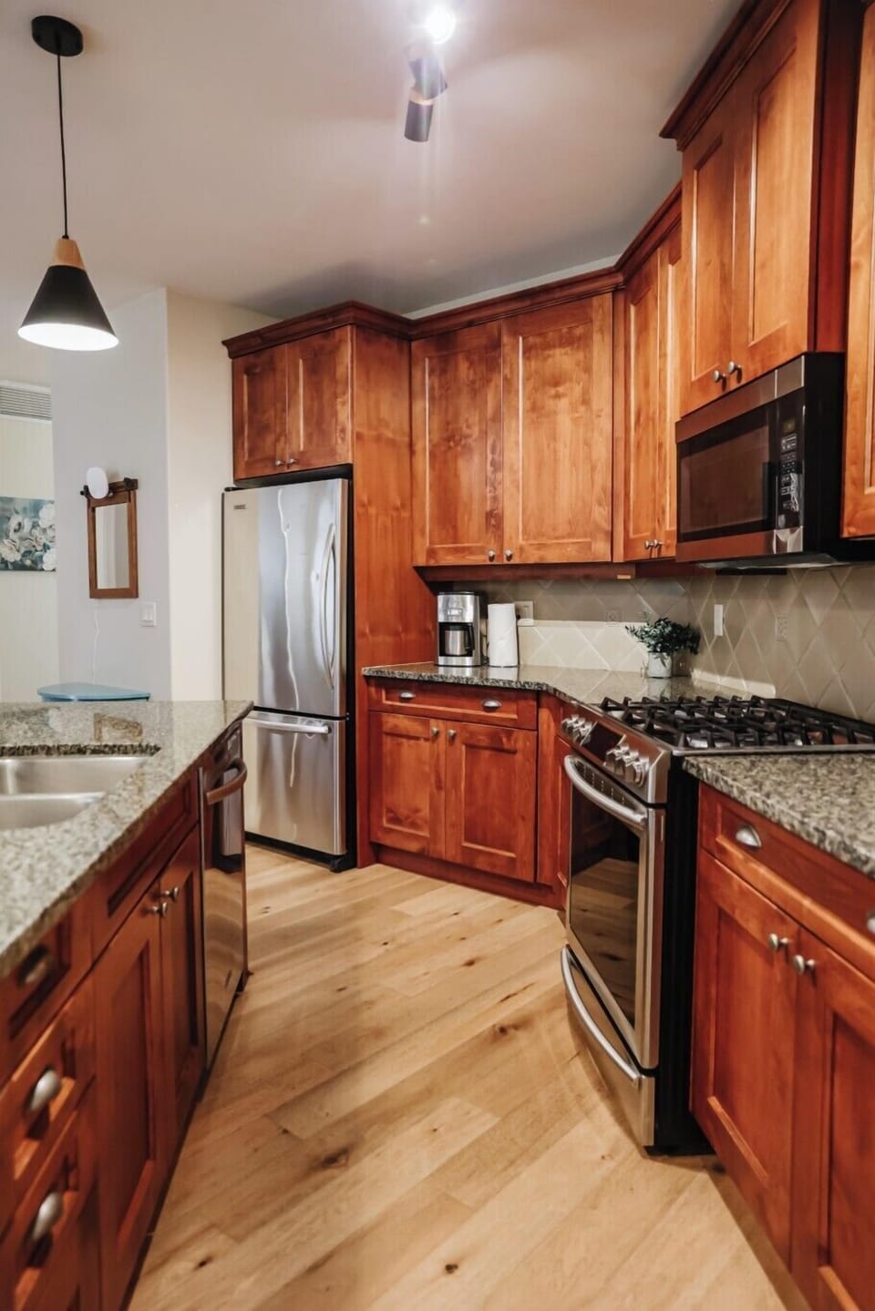 This bright kitchen features warm wooden cabinetry, stainless steel appliances, and dark granite countertops. A coffee maker sits on the counter, with a stove and built-in microwave, all illuminated by a stylish black pendant light.