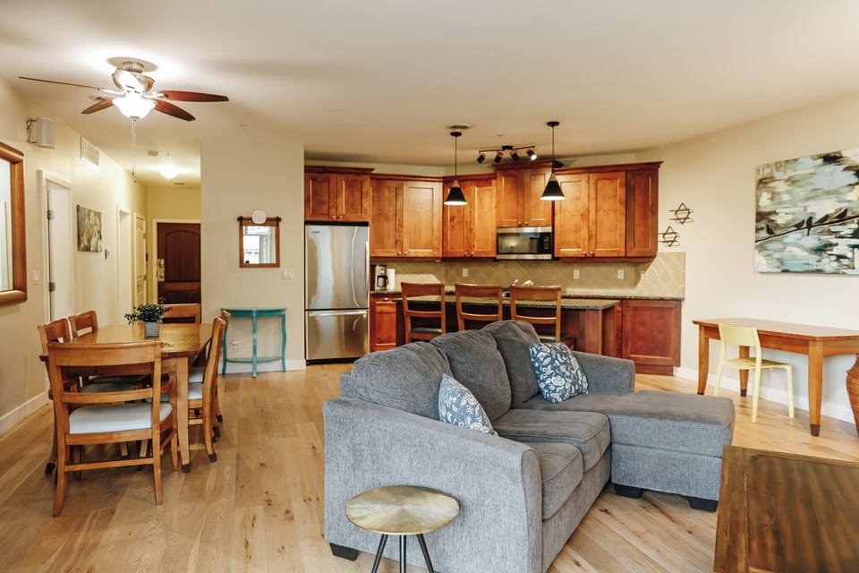 A stylish living area with a gray sectional sofa, patterned pillows, and a wooden coffee table. The kitchen features sleek stainless steel appliances and a breakfast bar with four chairs, all on light wood floors.