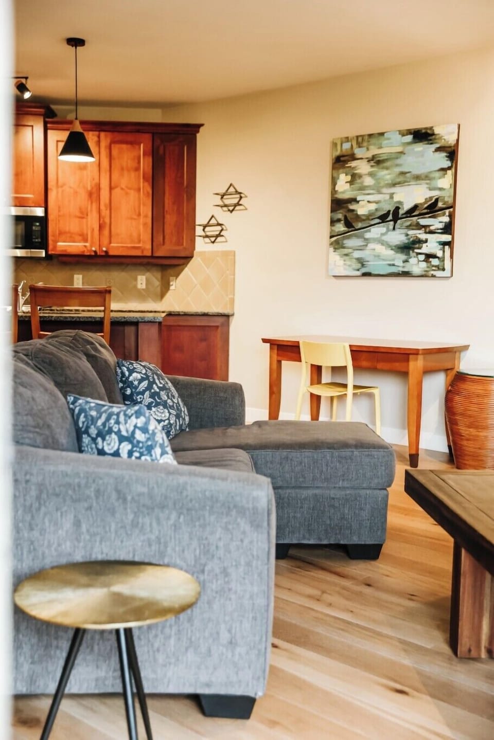 This inviting living room features a gray sectional sofa, a wooden coffee table, and a yellow-accented desk. Hardwood floors complement the space, with a large painting on the wall and a glimpse of the kitchen with wooden cabinetry.