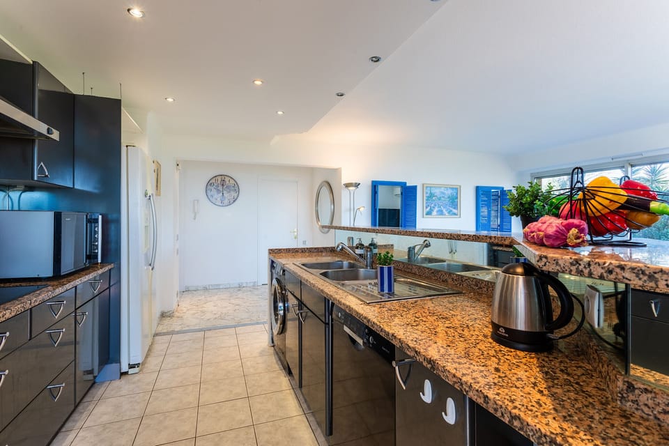 Kitchen connected to the living room with black cabinets, granite countertop, extractor fan, stainless steel sink, and modern appliances.