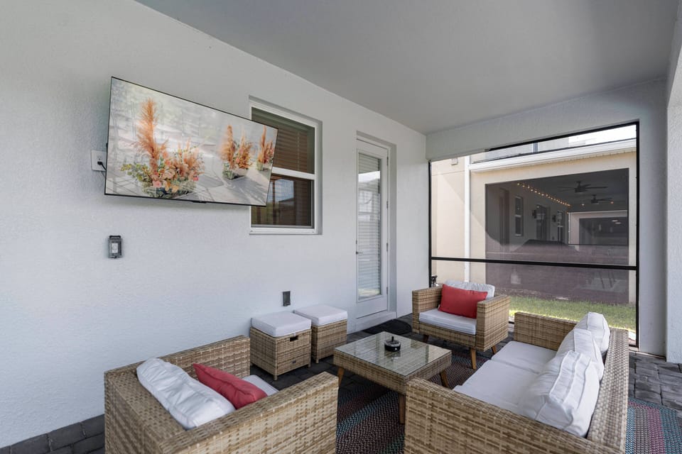 Relax in the cozy outdoor lounge area featuring stylish wicker furniture with plush white cushions and vibrant red accents. A large flat-screen TV offers entertainment as you enjoy the gentle breeze through the screened enclosure.