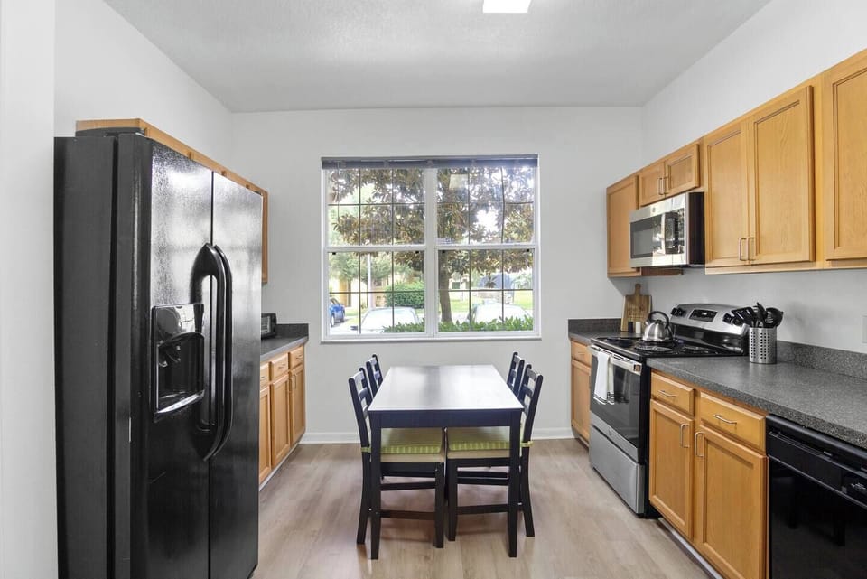 The kitchen features cabinets, a four-burner stove with an oven, a microwave, a kettle, a large refrigerator, and a dishwasher. It also includes a dining table for four, a bar area, and natural light that fills the space, creating a bright and welcoming atmosphere.