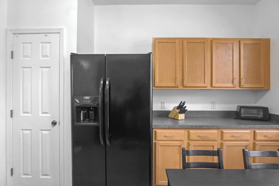 The kitchen features cabinets, a four-burner stove with an oven, a microwave, a kettle, a large refrigerator, and a dishwasher. It also includes a dining table for four, a bar area, and natural light that fills the space, creating a bright and welcoming atmosphere.