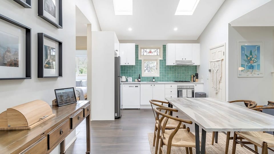 Skylit Dining & Tiled Charm - A bright, open-plan dining and kitchen area illuminated by overhead skylights, featuring a long marble-patterned table with woven chairs and a vibrant seafoam-green tiled backsplash.