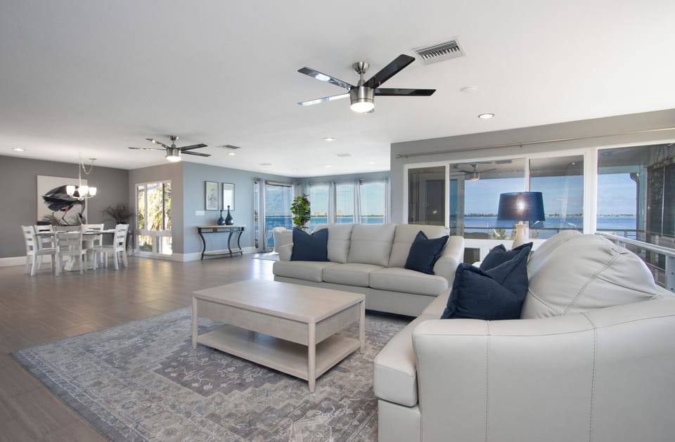 Bright, spacious living area with a leather sectional, dining space, and stunning waterfront views through large windows.