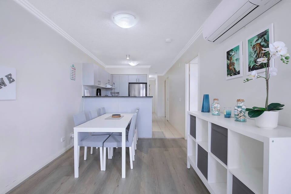 The dining area is a simple six-seater setting with a classic white wooden table and matching soft grey high-back chairs. 
