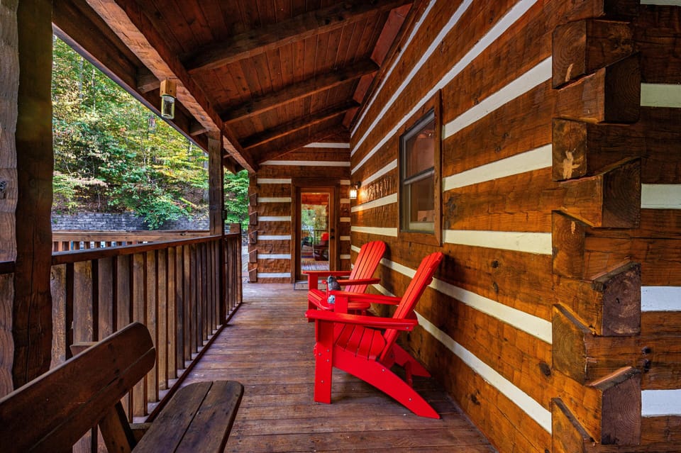 Wooden porch with rich log cabin walls, wide plank flooring, bright red Adirondack chairs, and rustic bench seating.