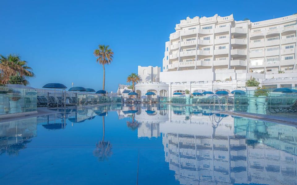 Reflective waters of a tranquil pool bordered by palm trees and a clear blue sky, offering an urban oasis.