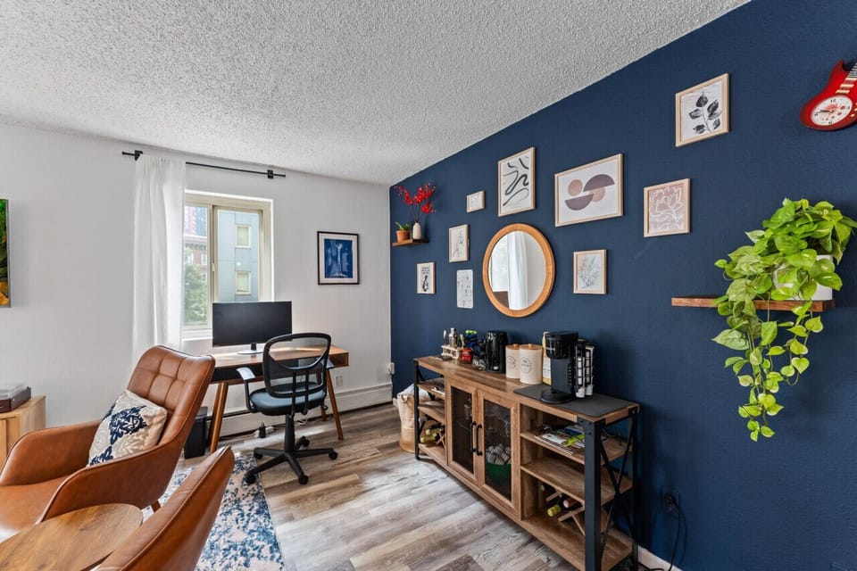 A dedicated desk with a monitor sits by a bold blue accent wall with stylish decor. The wooden console offers a coffee station and storage, with a touch of greenery adding charm.
