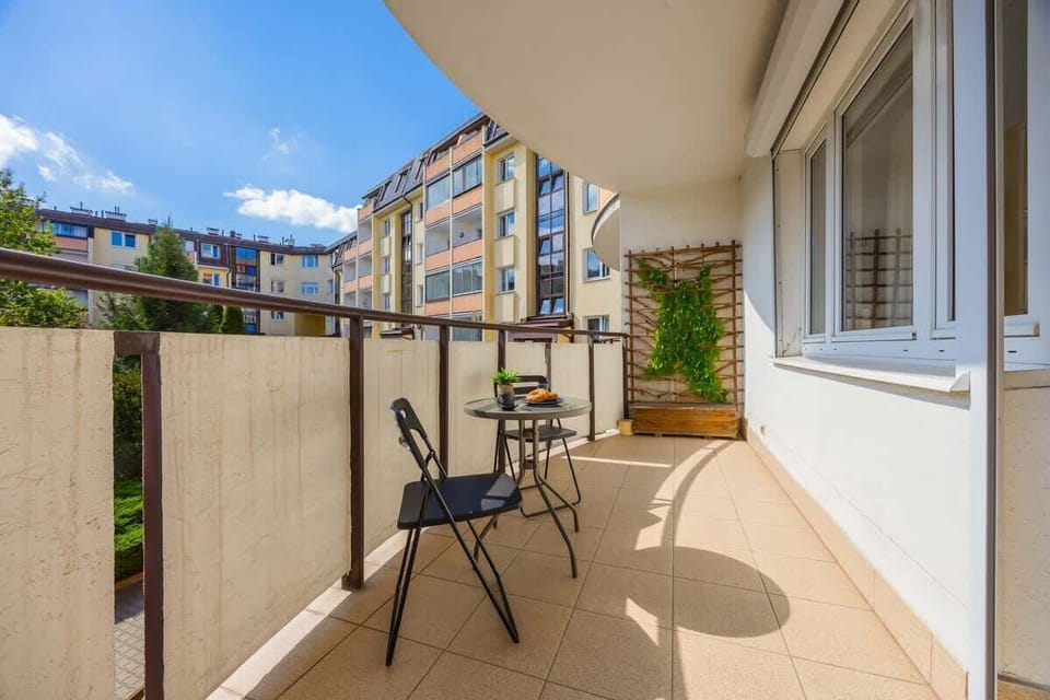 A cozy balcony with a table and two chairs, overlooking a street view with trees and neighboring buildings.