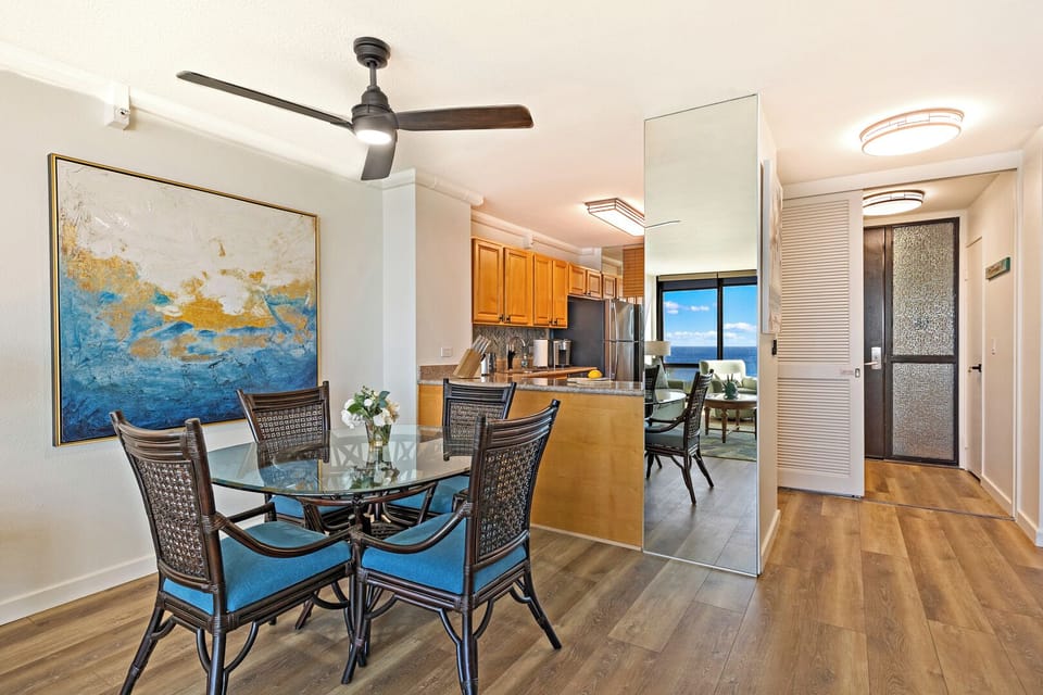 Mahana 718’s dining area features a glass-top table, modern fan, and vibrant artwork.